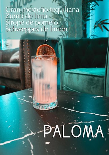 Cóctel Paloma con rodaja de naranja en una mesa de mármol. Fondo con plantas y sofá.